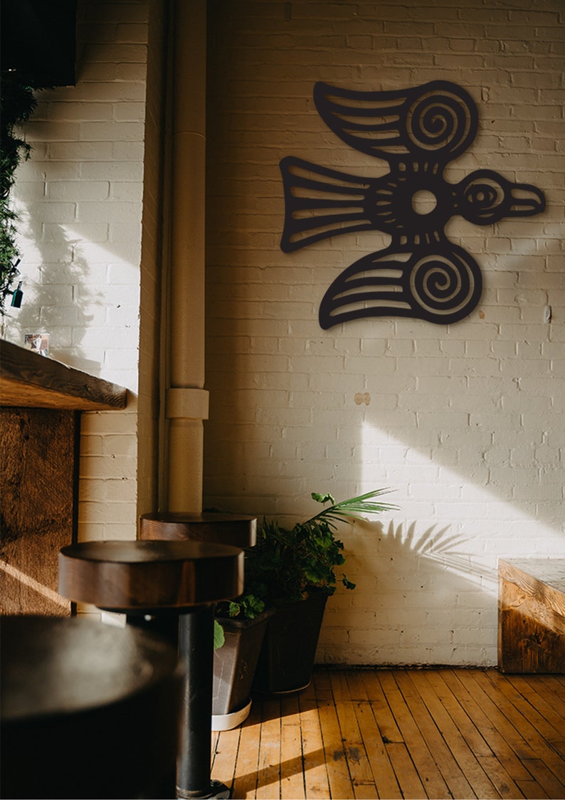 May include: A black metal wall art piece depicting a stylized bird with intricate details. The bird is flying with its wings spread wide and has a circular design in the center of its body. The art piece is mounted on a white brick wall.