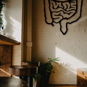 May include: A black metal cutout of a human intestine hangs on a white brick wall in a cafe setting. Two wooden bar stools are in the foreground, with a potted plant to the right.
