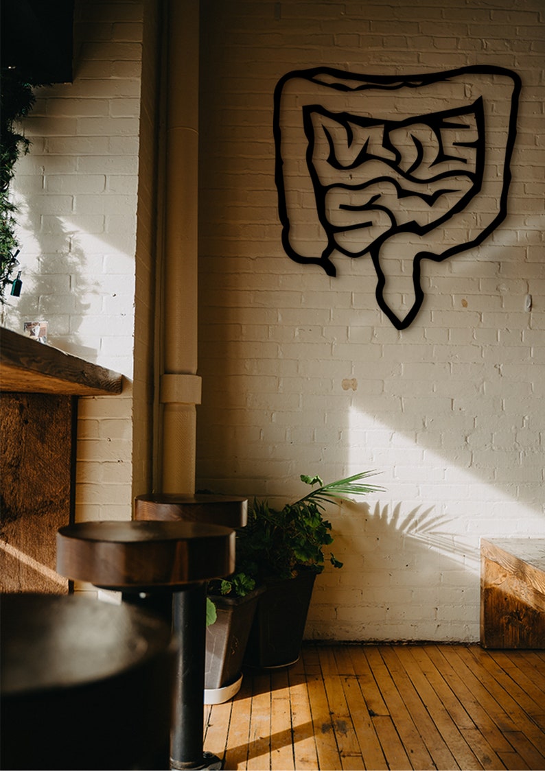 May include: A black metal cutout of a human intestine hangs on a white brick wall in a cafe setting. Two wooden bar stools are in the foreground, with a potted plant to the right.