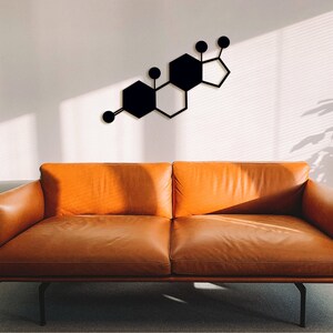 Puede incluir: Una escultura de pared de metal negro en forma de molécula química, con formas hexagonales conectadas por pequeños círculos, colgada en una pared blanca sobre un sofá de cuero marrón.