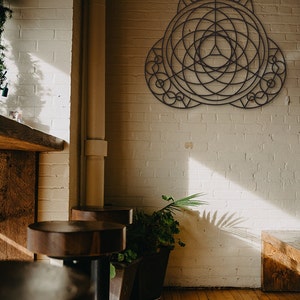 Puede incluir: Una pieza de arte de pared geom&eacute;trica de metal negro con un dise&ntilde;o circular, colgada en una pared de ladrillo blanco en una habitaci&oacute;n con pisos de madera y dos taburetes de bar marrones.