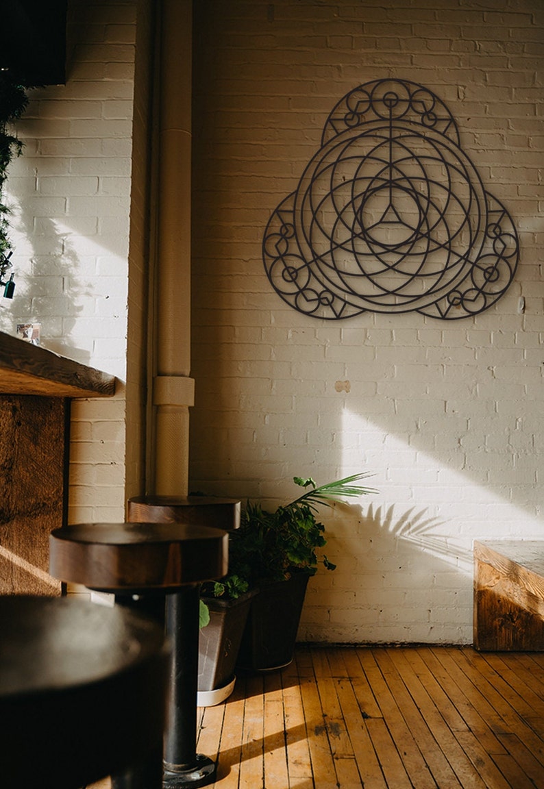 Puede incluir: Una pieza de arte de pared geom&eacute;trica de metal negro con un dise&ntilde;o circular, colgada en una pared de ladrillo blanco en una habitaci&oacute;n con pisos de madera y dos taburetes de bar marrones.