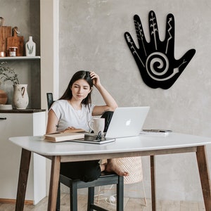 May include: A black metal wall decor in the shape of a hand with a spiral design and various lines and shapes inside. The decor is hanging on a gray wall in a room with a white table and a person sitting at the table.