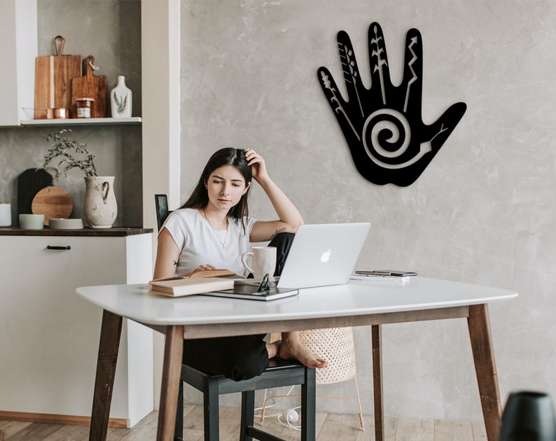 May include: A black metal wall decor in the shape of a hand with a spiral design and various lines and shapes inside. The decor is hanging on a gray wall in a room with a white table and a person sitting at the table.