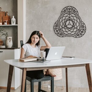 Puede incluir: Una mujer se sienta en una mesa blanca con un port&aacute;til, un libro y una taza. Una pieza de arte de pared geom&eacute;trica negra cuelga en la pared detr&aacute;s de ella.