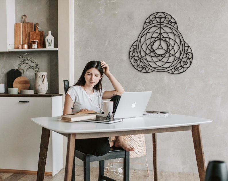 Puede incluir: Una mujer se sienta en una mesa blanca con un port&aacute;til, un libro y una taza. Una pieza de arte de pared geom&eacute;trica negra cuelga en la pared detr&aacute;s de ella.