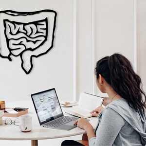 May include: A person sits at a white table with a laptop, a book, and a mug. A black outline of the human digestive system is on the wall behind them.