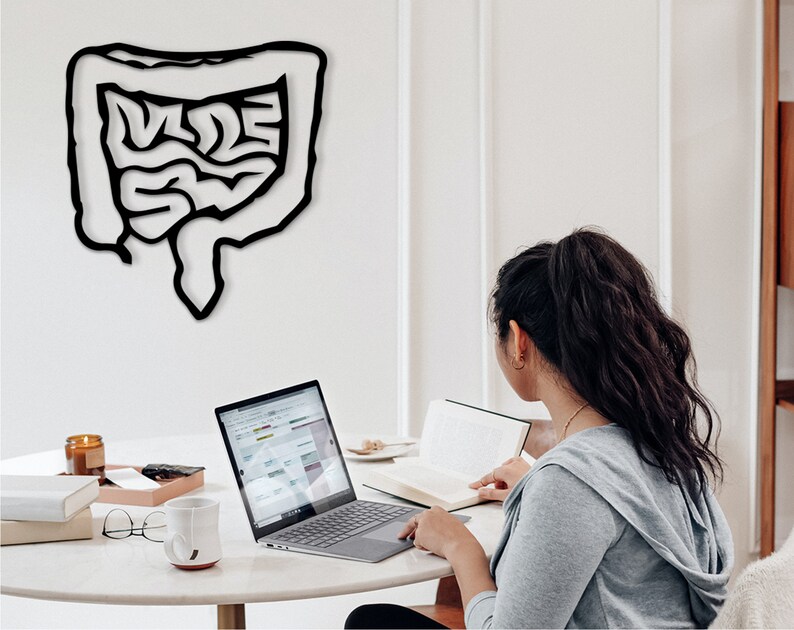 May include: A person sits at a white table with a laptop, a book, and a mug. A black outline of the human digestive system is on the wall behind them.