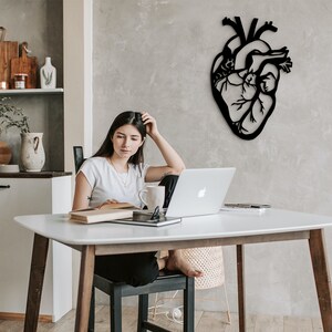 Op de afbeelding: Een vrouw zit aan een witte tafel met een laptop, een boek en een mok. Een zwarte metalen hartvormige wanddecoratie hangt aan de muur achter haar.