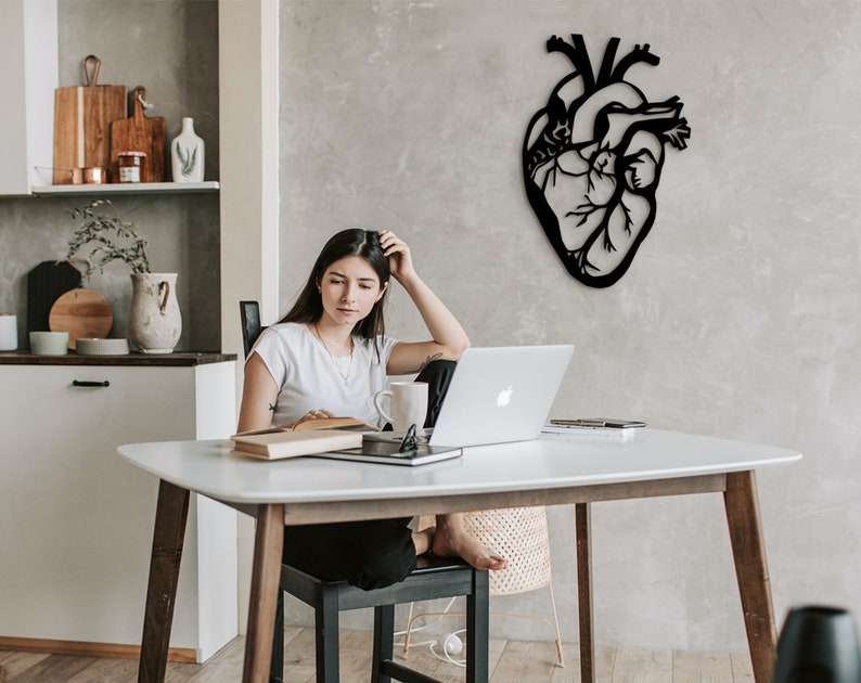 Op de afbeelding: Een vrouw zit aan een witte tafel met een laptop, een boek en een mok. Een zwarte metalen hartvormige wanddecoratie hangt aan de muur achter haar.