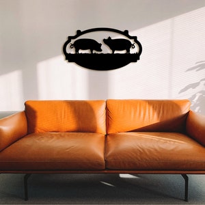 May include: Black metal wall decor featuring two pigs facing each other in an oval frame. The decor is mounted on a white wall above a brown leather sofa.