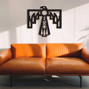 Puede incluir: Una escultura de pared de metal negro de un águila estilizada con patrones geométricos. El águila está montada en una pared blanca sobre un sofá de cuero marrón.