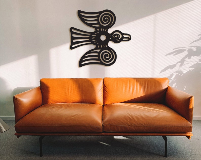 May include: A black metal wall art sculpture of a stylized bird with intricate details. The sculpture is mounted on a white wall above a brown leather couch.