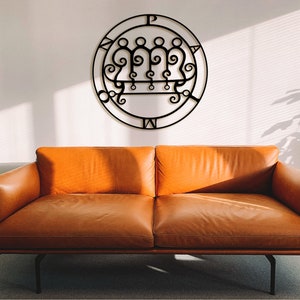 May include: A black metal wall art piece featuring a circular design with intricate symbols and text, mounted on a white wall above a brown leather sofa. The sofa has a modern design with a low profile and metal legs. The room is well-lit with natural light.