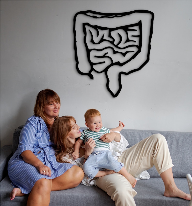 May include: Two adults and a child sitting on a gray couch. The adults are smiling and the child is looking at the camera. There is a black outline of a human intestine on the wall behind them.