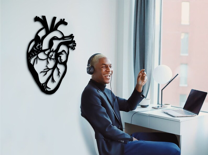 Op de afbeelding: Een zwarte silhouet van een anatomisch hart als wanddecoratie aan een witte muur. Een persoon met koptelefoon zit aan een bureau voor een raam.