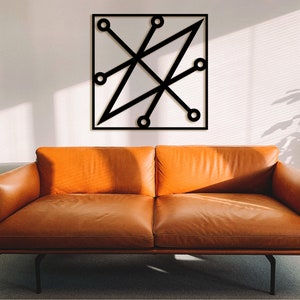 Op de afbeelding: Zwarte geometrische wandkunst met een vierkant frame, met elkaar kruisende lijnen en cirkelvormige accenten. Het kunstwerk is gemonteerd op een witte muur boven een oranje leren bank.