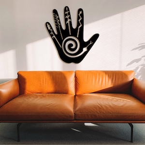May include: Black metal wall art in the shape of a hand with a spiral design and various lines and shapes on the fingers. The art is hanging on a white wall above a brown leather couch.