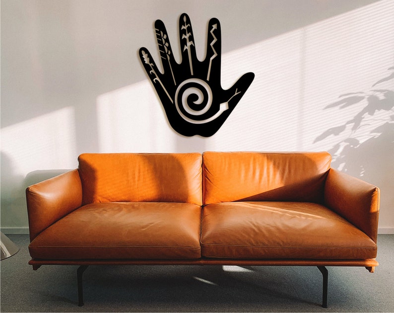 May include: Black metal wall art in the shape of a hand with a spiral design and various lines and shapes on the fingers. The art is hanging on a white wall above a brown leather couch.