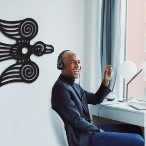 May include: A person wearing headphones sits at a desk in front of a laptop. The person is smiling and looking to the right. A black stylized bird design is on the wall behind the person.