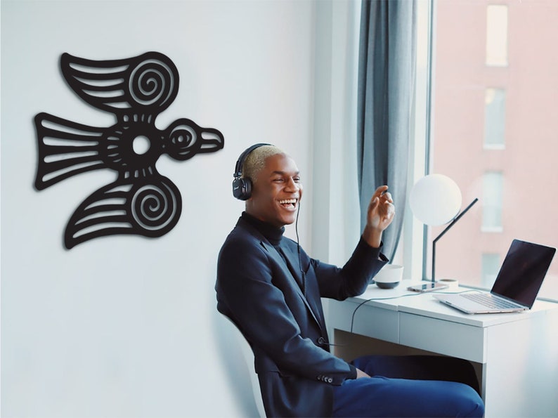 May include: A person wearing headphones sits at a desk in front of a laptop. The person is smiling and looking to the right. A black stylized bird design is on the wall behind the person.