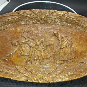 May include: A wooden serving tray with a carved design of people harvesting wheat. The tray is oval shaped and has a brown wood finish.