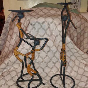 May include: A set of three metal candle holders in the shape of stylized figures. The figures are painted blue and have brown accents. The candle holders are on a patterned fabric background.