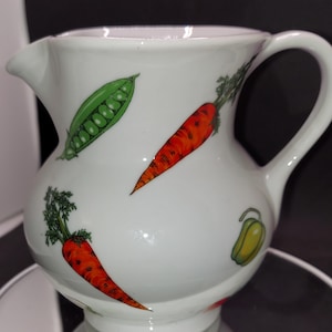 May include: White ceramic pitcher with a handle, featuring a colorful design of carrots, peas, and peppers.