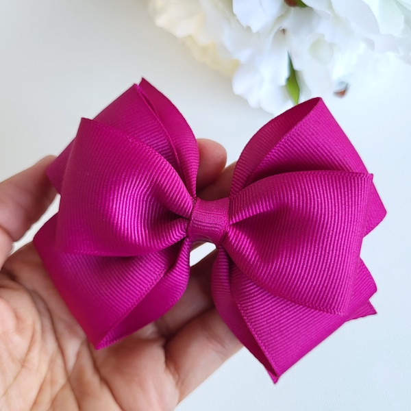 Puede incluir: Un lazo para el cabello de color fucsia vibrante hecho de cinta de grosgrain acanalada. El accesorio presenta un diseño estructurado de varias capas con bucles simétricos y un nudo central, mostrando un color rosa intenso y saturado.