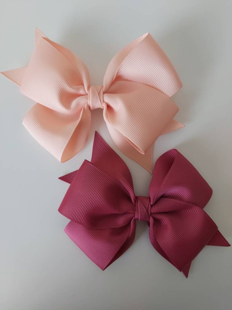 May include: Two hair bows, one peach and one burgundy, made of grosgrain ribbon. The bows are tied with a knot in the centre and have a pointed shape at the ends.