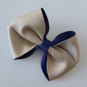 May include: A hair bow constructed from layered grosgrain ribbon in beige and navy blue. The design features a central navy blue knot that secures the loops of the beige ribbon, which is backed by a navy blue ribbon layer for a dual-tone effect.