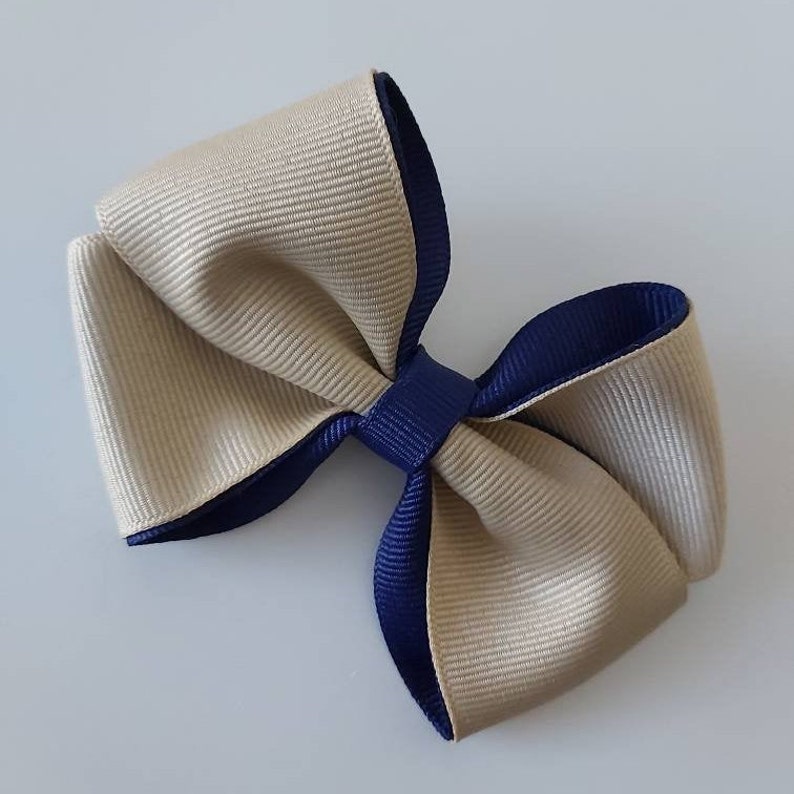 May include: A hair bow constructed from layered grosgrain ribbon in beige and navy blue. The design features a central navy blue knot that secures the loops of the beige ribbon, which is backed by a navy blue ribbon layer for a dual-tone effect.