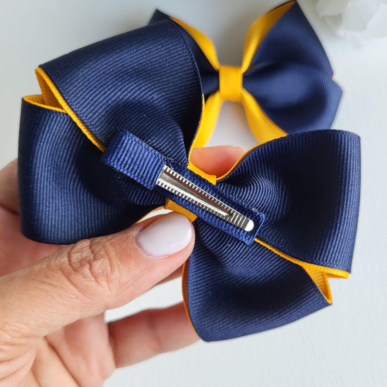 May include: Two layered hair bows made of navy blue and yellow grosgrain ribbon. The bows feature a stacked design with a navy blue center knot and a silver-toned metal alligator clip attached to the back of the primary bow for secure attachment.