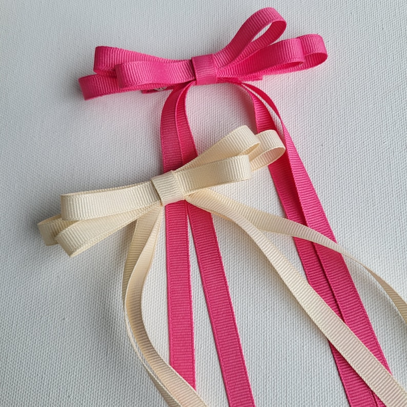 May include: Two hair bows, one pink and one cream, with long tails. The bows are tied together with a single knot.
