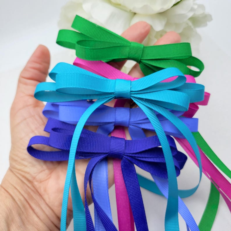 May include: Five colourful satin ribbon bows in shades of green, pink, blue, light blue, and dark blue. The bows are tied in a simple knot and the ribbons are long and flowing.