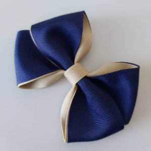 May include: A double-layered hair bow constructed from grosgrain ribbon. The bow features a deep navy blue outer layer and a contrasting beige inner layer, secured in the center with a matching beige ribbon knot. The texture is ribbed throughout.