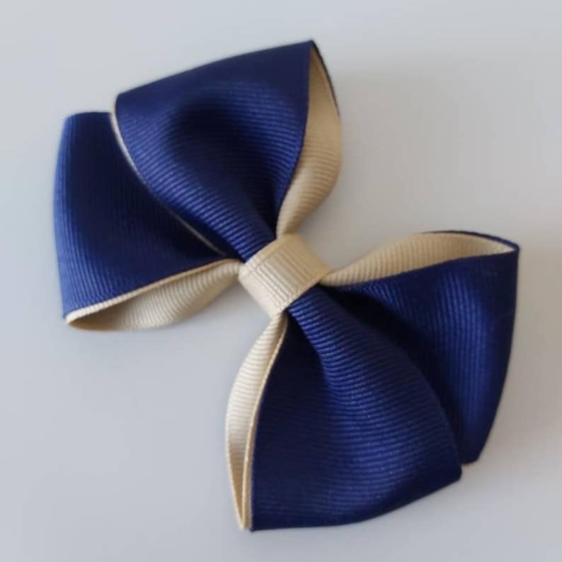 May include: A double-layered hair bow constructed from grosgrain ribbon. The bow features a deep navy blue outer layer and a contrasting beige inner layer, secured in the center with a matching beige ribbon knot. The texture is ribbed throughout.