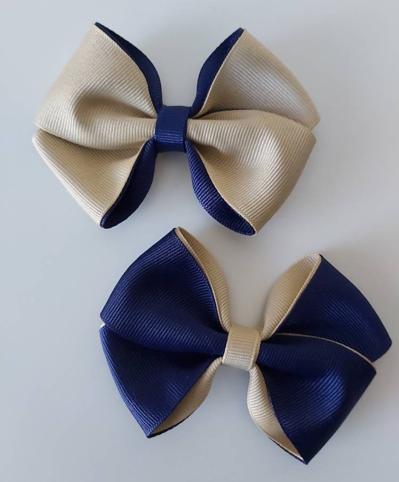 May include: Two hair bows made of ribbed grosgrain ribbon. The top bow is primarily beige with navy blue edges and a navy blue center knot. The bottom bow is primarily navy blue with beige edges and a beige center knot. Both bows feature a layered design.