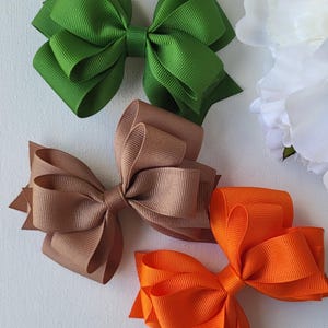 May include: Three colorful hair bows in shades of green, brown, and orange. The bows are made of a ribbed fabric and are arranged on a white background. The bows are a classic accessory for various hairstyles.