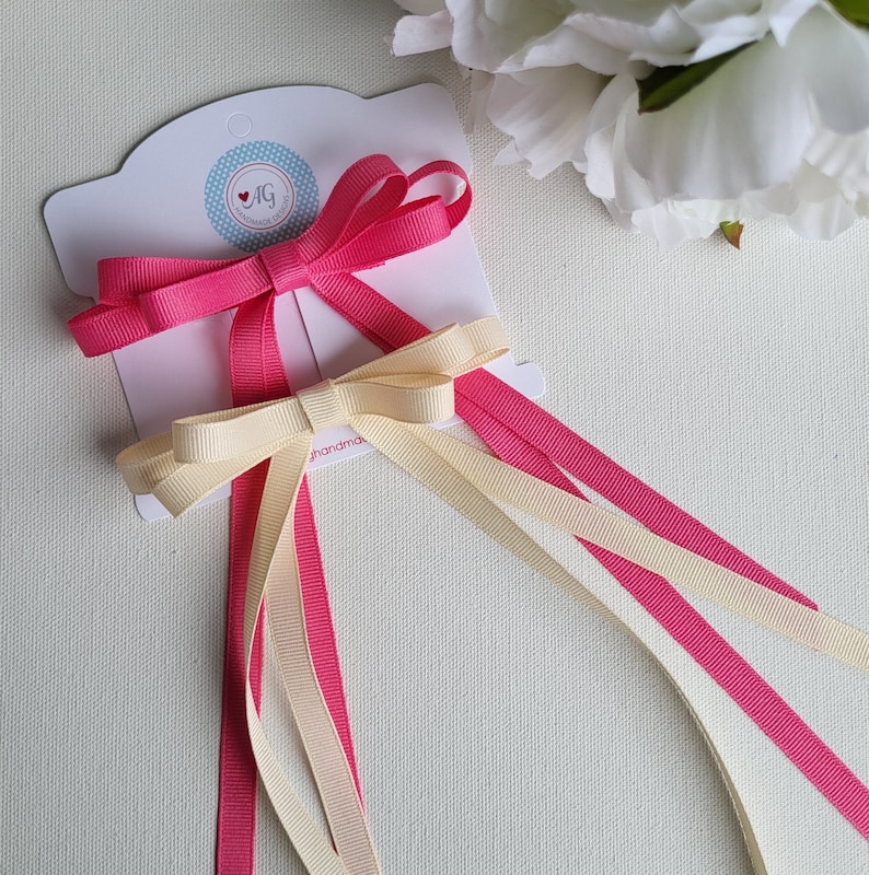 May include: Two hair bows with long tails. One bow is pink and the other is cream. The bows are on a white card with the text "AG Handmade Goods" printed on it.