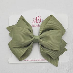 May include: A sage green hair bow made of ribbed grosgrain ribbon, featuring a multi-layered design with a central knot. The bow is attached to a white display card with the text AG handmade and the website www.shopaghandmadedesigns.