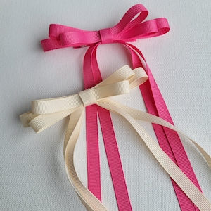 May include: Two hair bows, one pink and one cream, with long tails. The bows are tied together with a single knot.