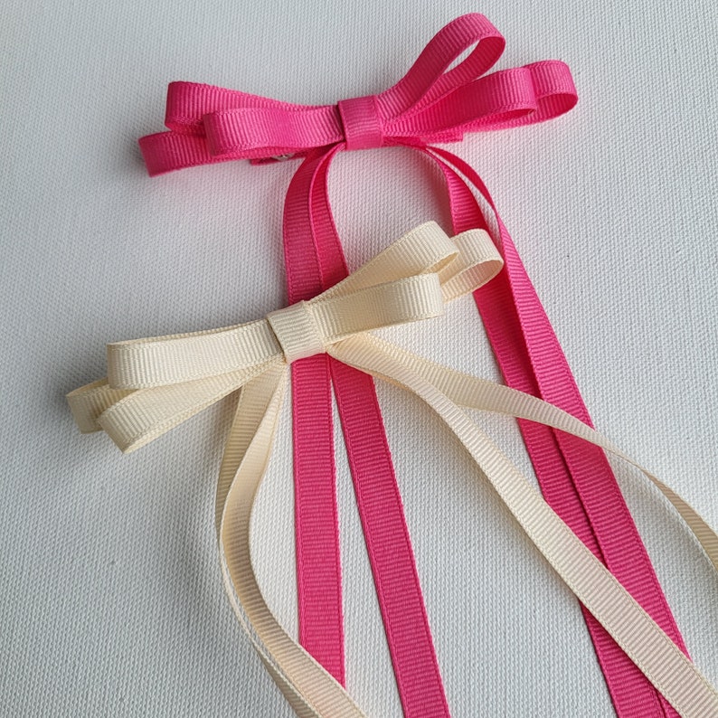 May include: Two hair bows, one pink and one cream, with long tails. The bows are tied together with a single knot.