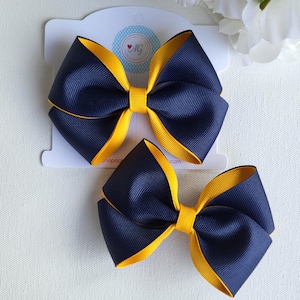 May include: Two layered hair bows made of navy blue and bright yellow grosgrain ribbon. The bows feature a stacked design with a central knot. The top bow is attached to a white display card with a circular logo containing the letters AG.
