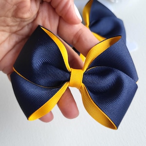 May include: A layered hair bow constructed from navy blue and bright yellow grosgrain ribbon. The design features a wide navy blue ribbon base with a contrasting yellow ribbon border, secured in the center by a matching yellow ribbon knot.