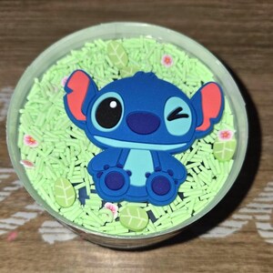 May include: A clear plastic container filled with green sprinkles and a blue cartoon character. The character has large ears, a winking eye, and is surrounded by small flower and leaf-shaped decorations.