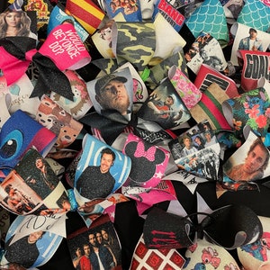 May include: A collection of colorful hair bows with various celebrity and pop culture themes. The bows feature images of singers, bands, and cartoon characters. Some of the bows have glitter and sequins.