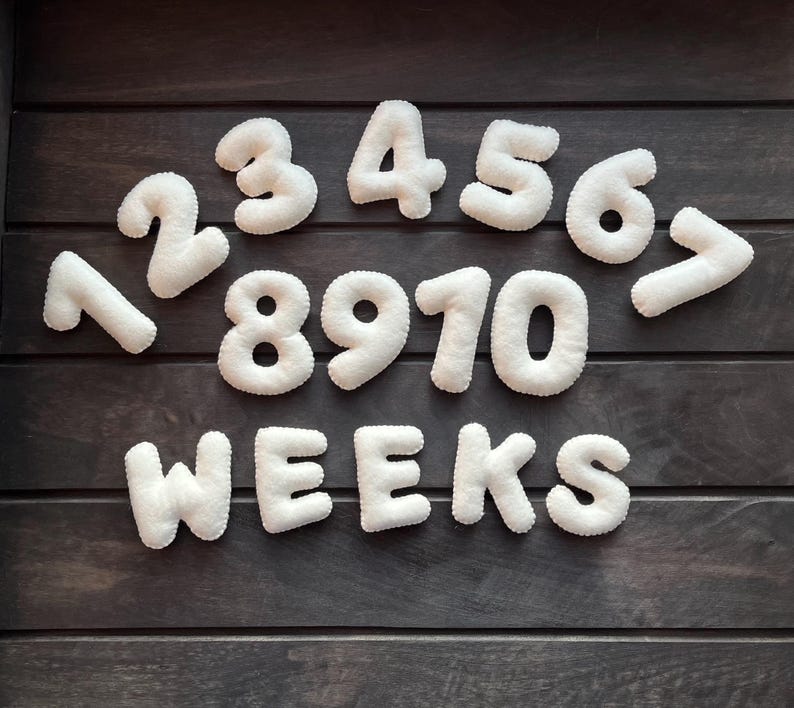 May include: White felt numbers 1 through 10 and the word "WEEKS" arranged in a semi-circle on a dark wood background.
