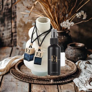 May include: A still life arrangement featuring a black spray bottle and two car air fresheners. The spray bottle has gold text that reads "Golden Noir" and "Essentially Infused Candle Co." The air fresheners are square with the letters "EI" on them.