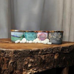 May include: Four scented candles with gold lids and decorative labels. The candles are arranged in a row on a wooden surface. The labels read "Fresh Fall Morning", "Mountain Teawood", "Harvest Pomegranate", and "Sweet Whiskey".